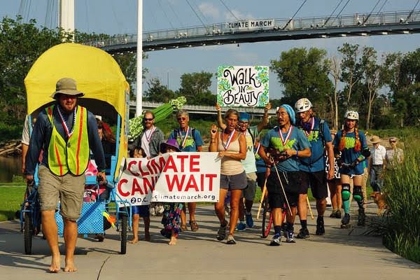 This was one wave of our group passing the Bob Kerrey Pedestrian Bridge which connects Omaha, Nebraska to Des Moine, Iowa, over the Missouri River. That was me in the front pulling a shaded cart trolley carrying two children. In this wave of walkers there are two signs held, one saying 'Walk in Beauty' and the other 'Climate can't wait.; One of the walkers is roller skating. The pedestrian bridge in the background having a second wave is hanging a banner which says climate march, visibly from several hundred feet away.